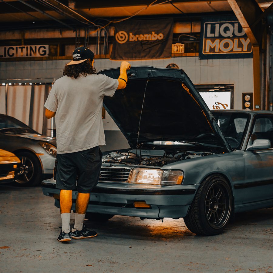 A man seen from behind, one hand on the raised hood of a grey Toyota Cressida, in a repair bay hung with Brembo and Liqui Moly banners