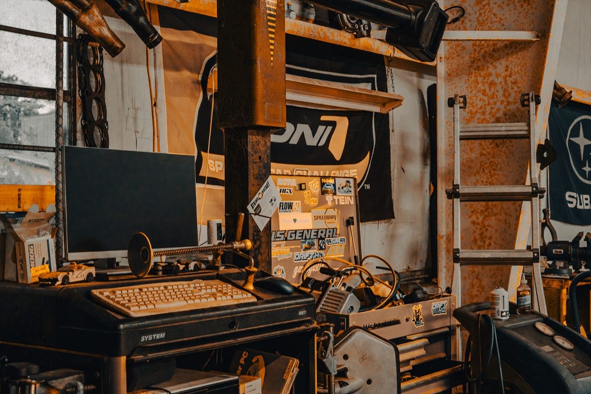 A CRT monitor and a grease-marked keyboard on a rolling cart in a repair bay, wedged between a lift post and a shelf of parts